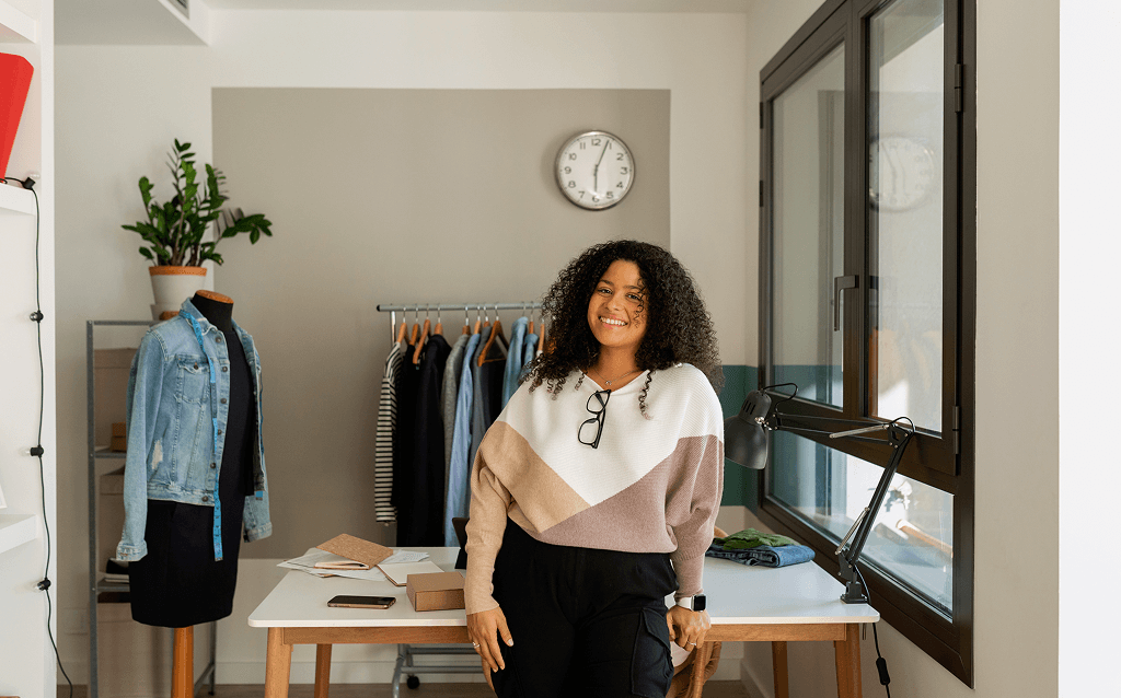Mulher jovem de cabelos cacheados e sorriso confiante em um ateliê de moda. Ela está apoiada em uma mesa com materiais de trabalho, enquanto um manequim com uma jaqueta jeans