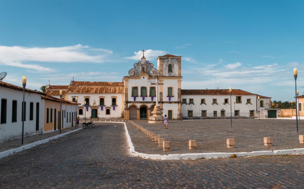 Imagem de uma praça histórica com um edifício colonial e céu azul. A arquitetura destaca elementos da cultura local e do patrimônio histórico.