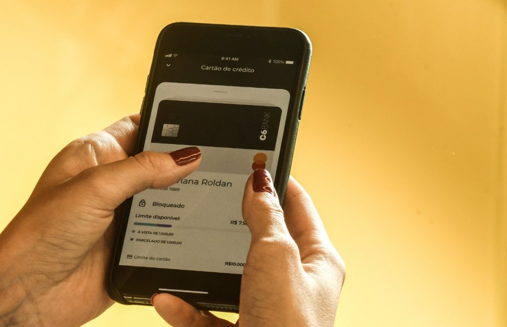 Foto das mãos de uma mulher branca, com as unhas pintadas de vinho, segurando um celular onde se pode ver o aplicativo do C6 Bank na tela.