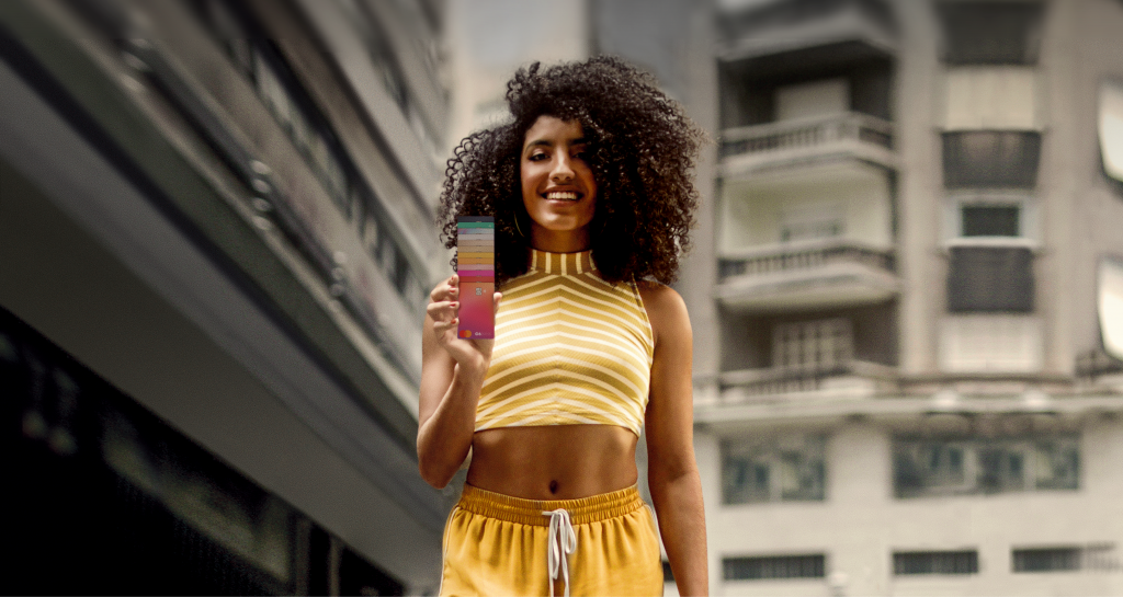 Foto de uma mulher negra, com cabelo afro, cropped dourado e shorts amarelo, sorrindo e segurando os cartões do C6 Bank em uma fileira vertical com a mão direita. Ao fundo, prédio do centro de São Paulo.