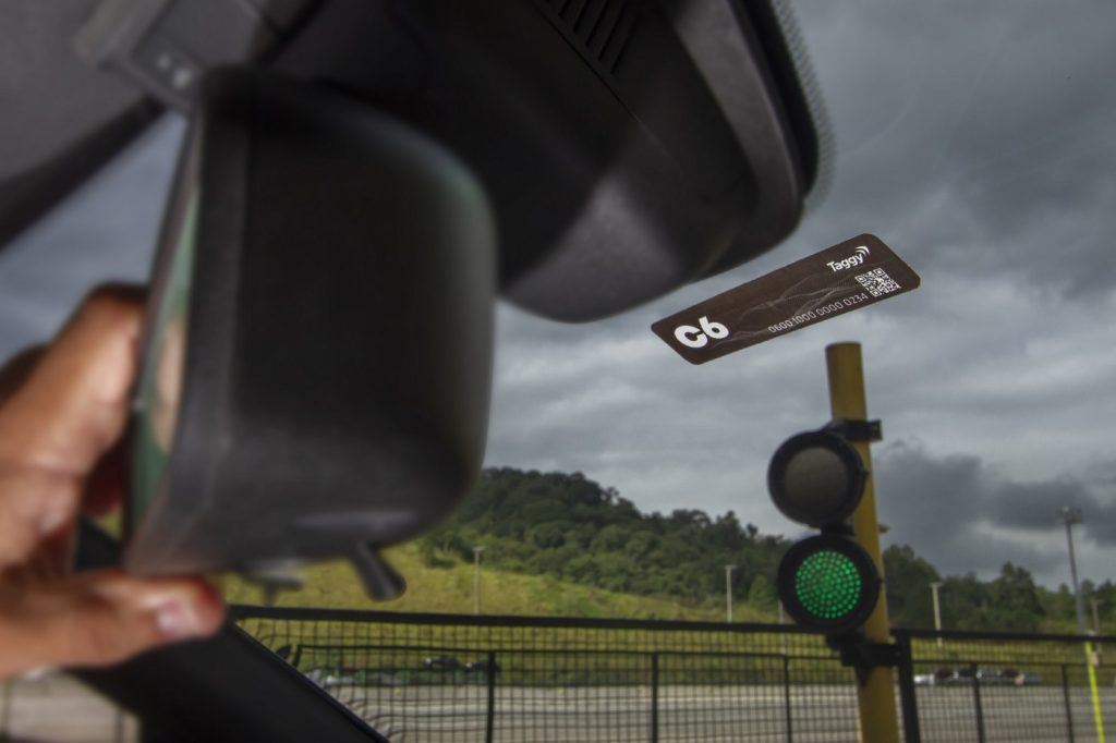 Foto do parabrisa de um carro com o adesivo do C6 Taggy colado um pouco abaixo do retrovisor. Ao fundo, o sinal verde do pedágio aberto.