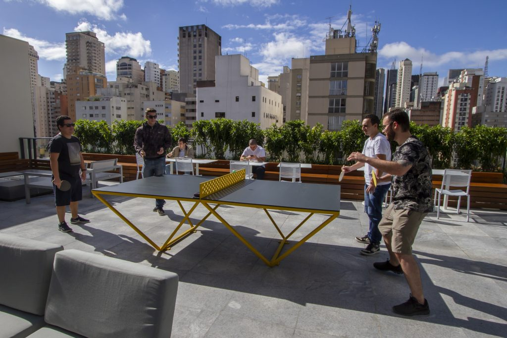 Foto da parte externa da área de descompressão no oitavo andar do prédio do banco. Quatro pessoas, sendo duas de cada lado, estão ao redor de uma mesa de pingue pongue preta, com redinha e bases amarelas. Ao fundo, podemos ver algumas mesas e cadeiras, alinhadas a um pequeno jardim na extremidade do prédio. Atrás dele, um skyline de prédio de São Paulo, de baixo de um céu azul com algumas poucas nuvens brancas.