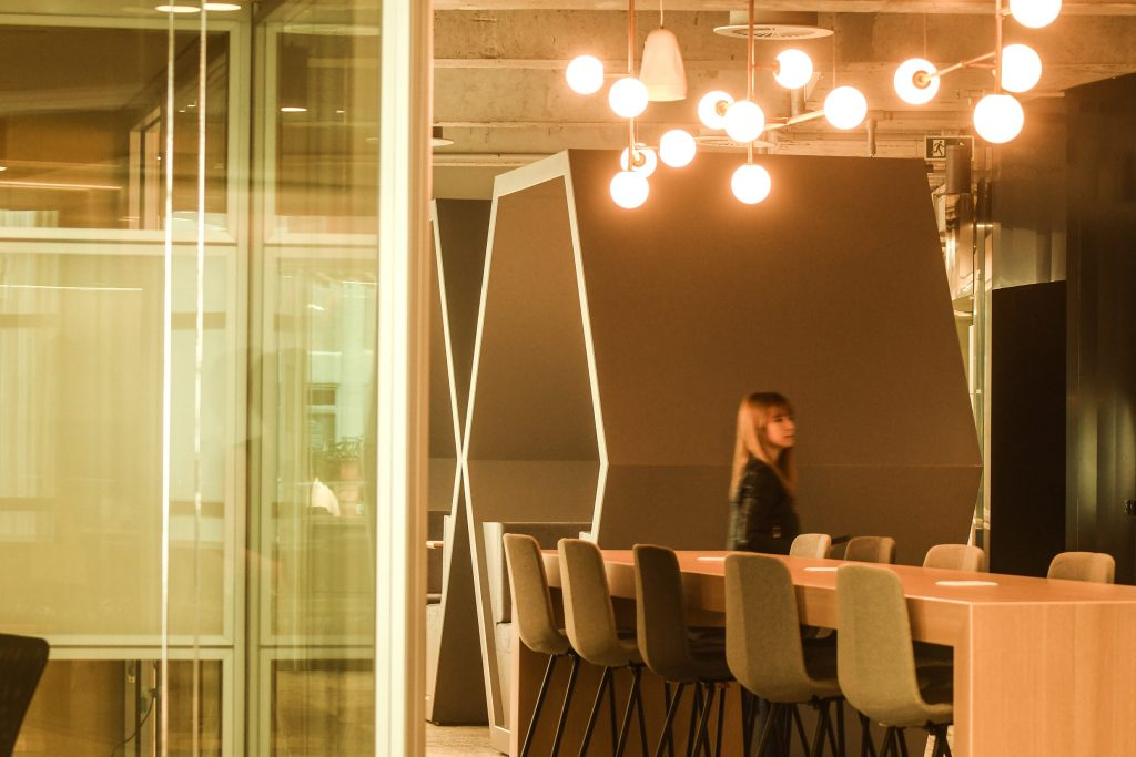 Foto de um ambiente interno do C6 Bank, que mostra uma longa mesa de madeira para refeições, com cadeiras altas colocadas dos dois lados, uma luminária moderna com luzes amarelas e uma grande estrutura em formato hexagonal, com dois bancos e uma mesa dentro. Na foto também é possível identificar uma mulher ao centro da foto, mas em segundo plano.