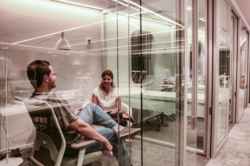 Foto de uma sala de reunião pequena no prédio do C6 Bank. Através das paredes de vidro, é possível ver um homem e uma mulher sentados em cadeiras de escritório, conversando. O carpete e a parede são cinzas. Ao fundo, é possível ver uma segunda sala de reunião, com uma janela em formato oval e um bancada com telefone.