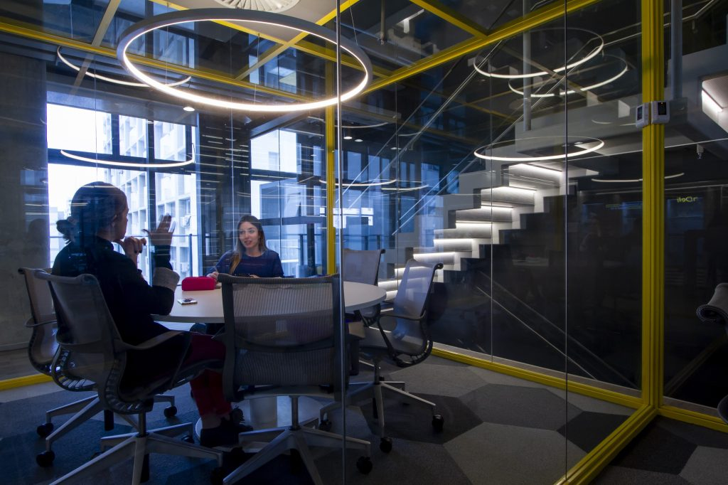 Foto de uma das salas de reunião do C6 Bank, onde se pode ver uma mesa circular no centro da sala e duas mulheres sentadas ao redor dela. As paredes da sala são de vidro, então também e possível ver uma escada com os degraus iluminados ao lado e uma varanda do prédio ao fundo.