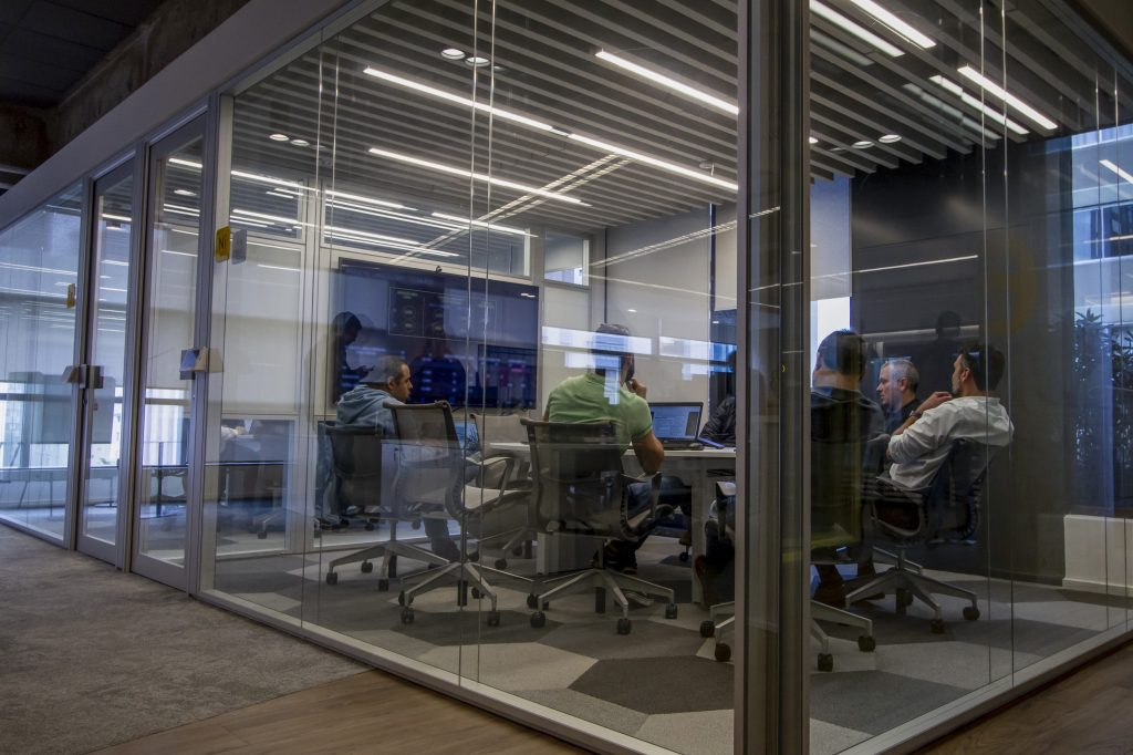 Foto de uma sala de reunião do prédio do C6 Bank. A sala tem paredes de vidro e, através dela, podemos ver uma mesa oval, cercada de pessoas sentadas em cadeiras de escritório ao redor, e uma televisão na parede. O carpete tem formatos hexagonais em diferentes tons de cinza e a iluminação no teto é indireta.