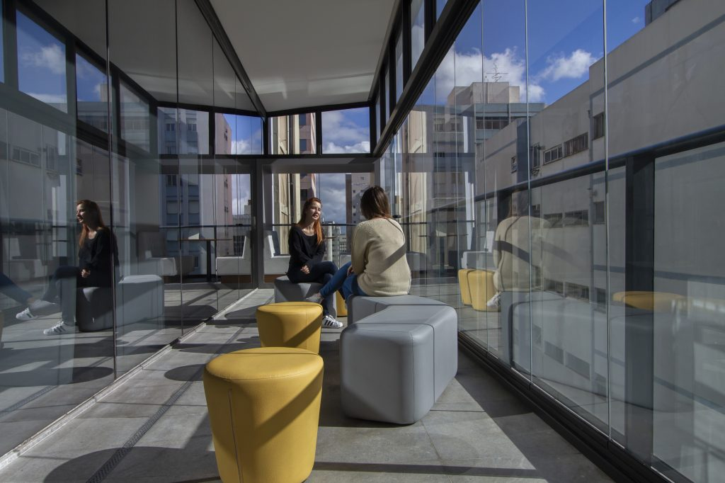 Foto de uma varanda do prédio do C6 Bank, onde é possível ver duas mulheres sentadas em pufs coloridos, alguns amarelos, outros cinzas. As janelas da varanda são totalmente de virdro, permitindo ver um prédio ao lado e o céu azul com poucas nuvens brancas ao fundo.