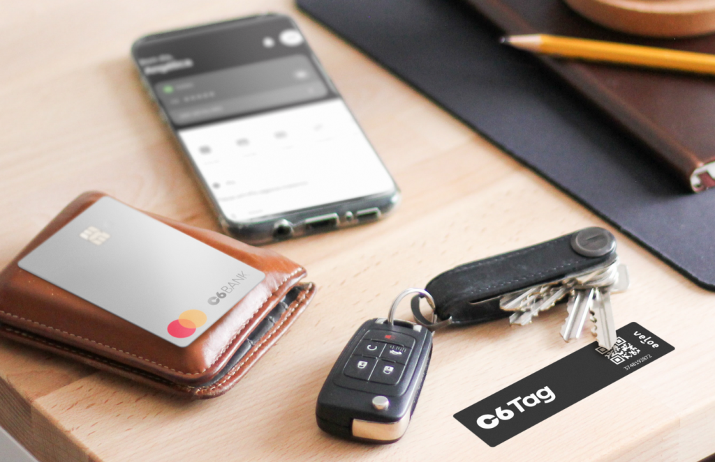 Foto de uma mesa onde é possível ver um celular com o aplicativo do C6 Bank aberto na tela, uma carteira com um cartão do C6 Bank apoiado em cima, a chave de um carro e uma C6 Tag