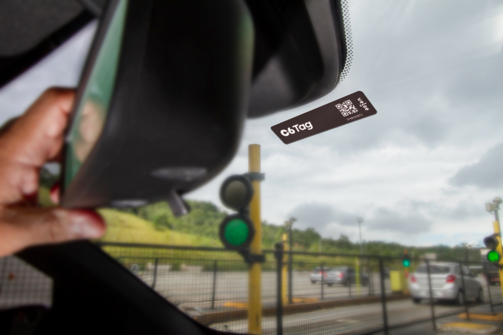 Foto do parabrisa de um carro que está passando por um pedágio, onde se pode ver o semáforo verde em segundo plano e o C6 Tag colado no parabrisa atrás do retrovisor em primeiro plano