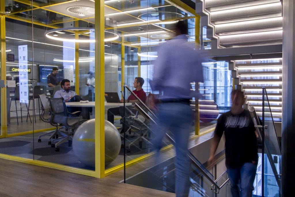 Foto de uma sala de reunião do prédiod o C6 Bank, ao lado de uma escada. Na foto, é possível ver duas pessoas dentro da sala, duas pessoas subindo ou descendo a escada e, ao fundo, através das paredes de vidro da sala, vemos o Carbon Deli.