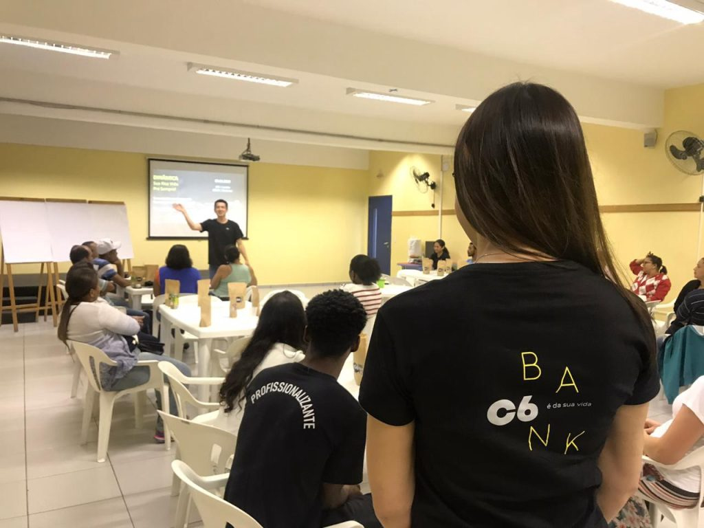 Foto de uma sala de aula, onde se pode ver o professor Liao ao fundo apontando para um telão, várias pessoas sentadas em frente a ele e uma pessoa de costas, em primeiro plano, com a camiseta do C6 Bank.