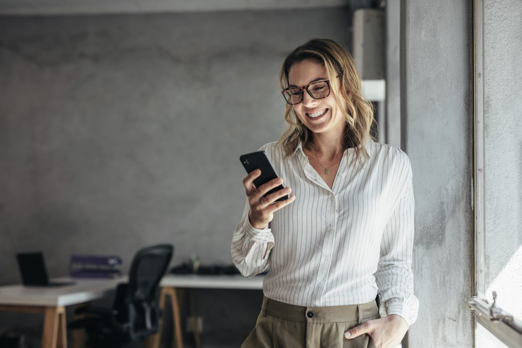 Mulher branca, de camisa e calça social, olhando para o celular próximo de uma janela, pesquisando sobre o Samsung Rewards