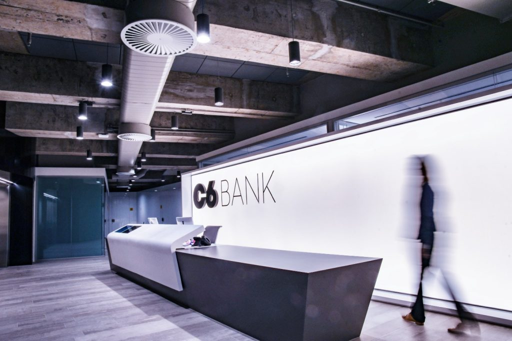 Foto da recepção do 7º andar do prédio do C6 Bank. Na foto consta a longa bancada da recepção, em frente a um painel com o logo do C6 Bank. Entre eles, caminha uma mulher.