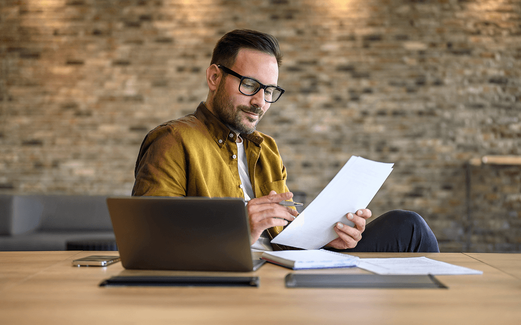 Homem com óculos e camisa mostarda analisa documentos em frente ao notebook, sentado à mesa em ambiente corporativo moderno.