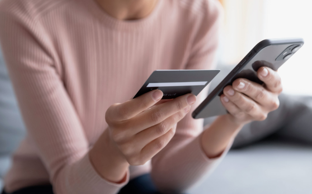 Pessoa realizando uma transação online segura, segurando um cartão de crédito na mão e utilizando um smartphone. A imagem retrata a modernidade das compras digitais.