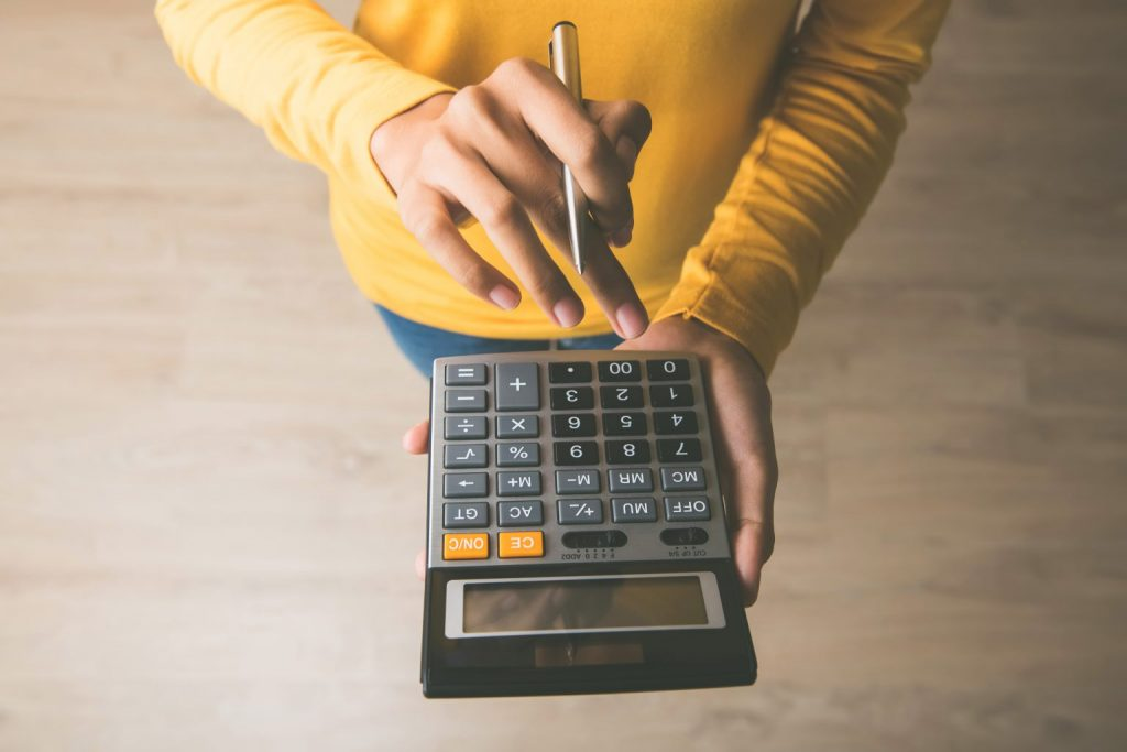 Pessoa de blusa de manga longa amarela, segurando uma calculadora em uma mão e uma caneta na outra, calculando o salário líquido