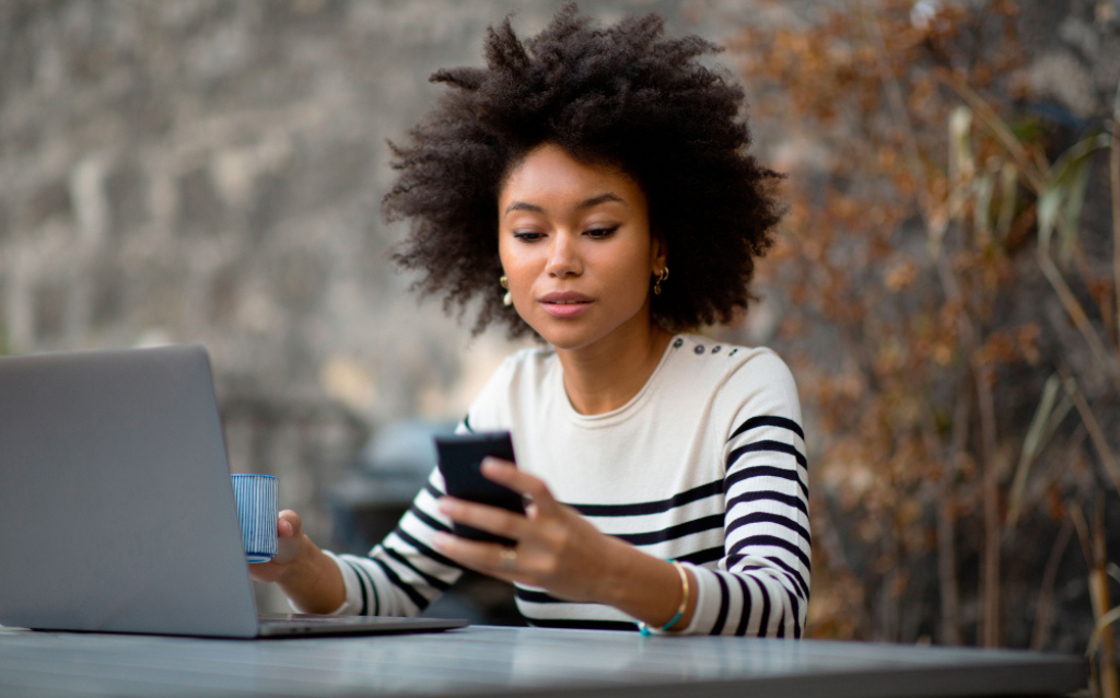 Mulher negra sentada em frente ao computador olhando para o celular e pensando em como deixá-lo mais seguro