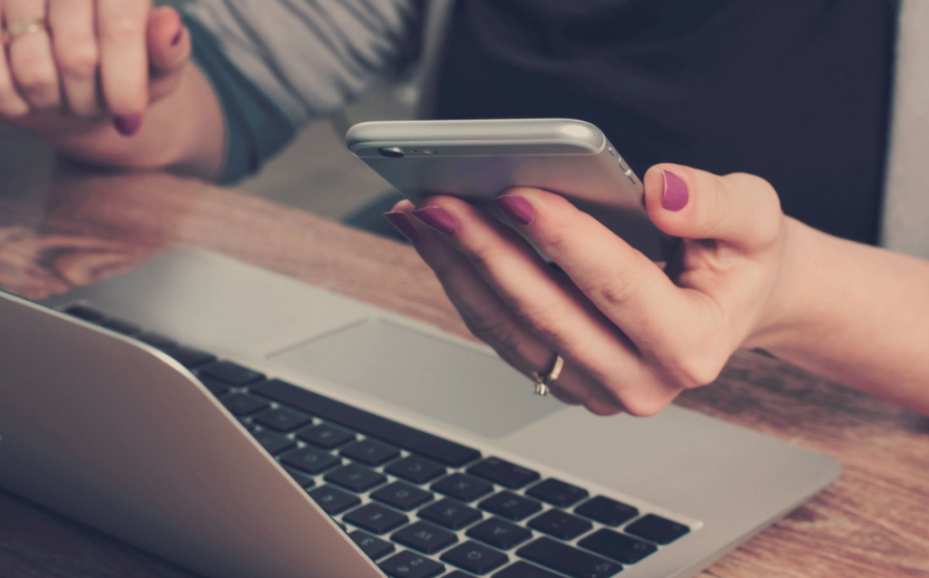 Uma mão feminina segurando um celular em frente ao computador enquanto pesquisa como evitar chargeback