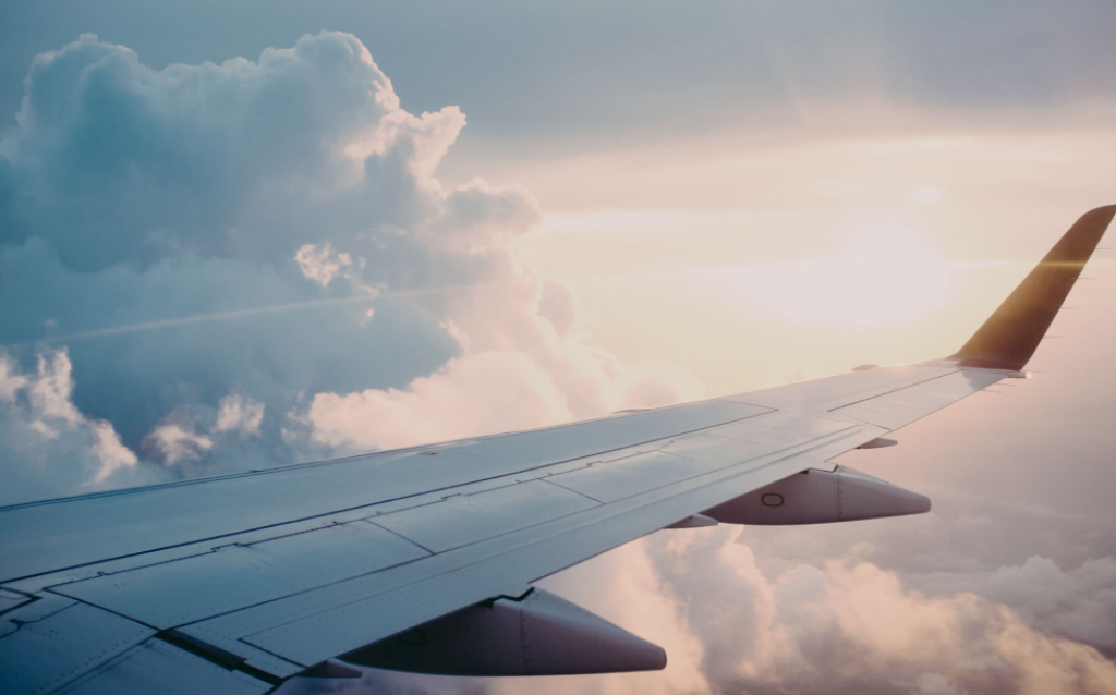 Asa de avião em voo, cercada por nuvens e sol, simbolizando organização para viagens.
