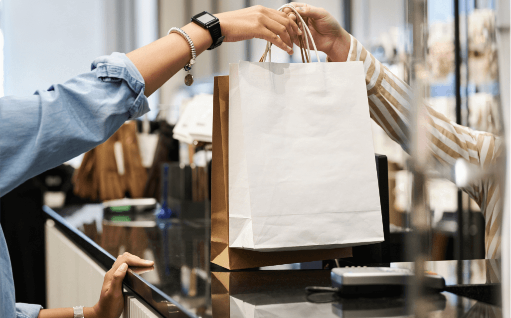 Imagem de uma pessoa recebendo sacolas de papel em um balcão de loja, representando uma compra concluída. O ambiente é moderno e organizado, com destaque para a interação entre o cliente e o atendente. A cena simboliza a importância de desenvolver uma relação consciente e equilibrada com o consumo.