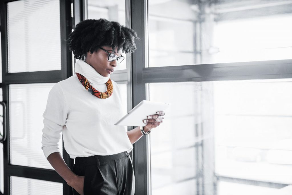 Mulher negra próxima à janela pesquisando no tablet o estágio da declaração do IR 2022