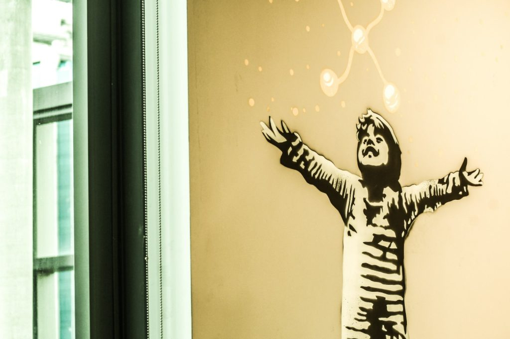 Foto de uma grafite em uma parede interna do prédio do C6 Bank. O desenho é de uma criança sorrindo, olhando pra cima, com os braços aberto. Acima dela, luzes.