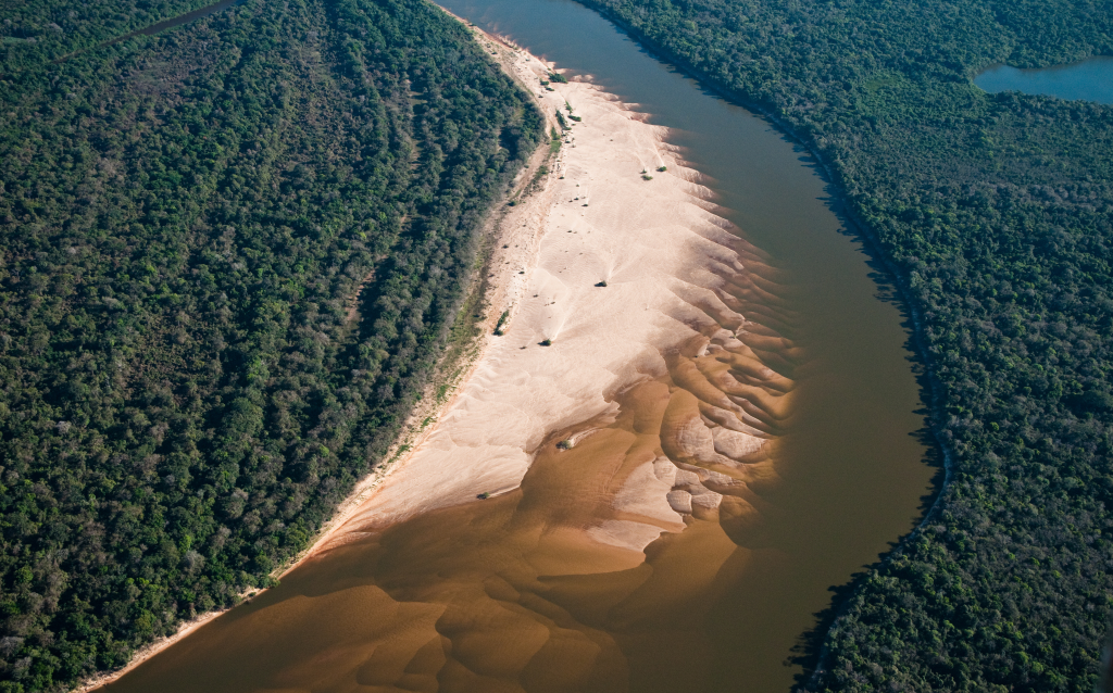 Vista aérea de um rio rodeado por vegetação exuberante, comoo destaque uma praia de areia clara e formações naturais na margem.