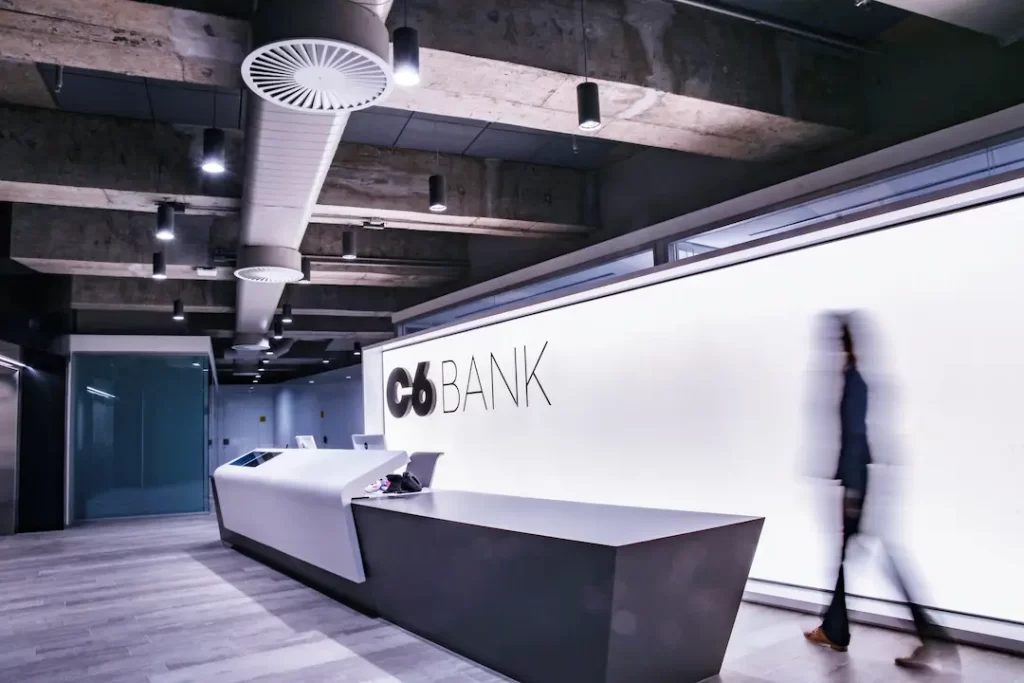 Imagem da recepção do C6 Bank com decoração industrial e tons claros com um vulto de uma pessoa passando pelo letreiro escrito &quot;C6 Bank&quot; em preto