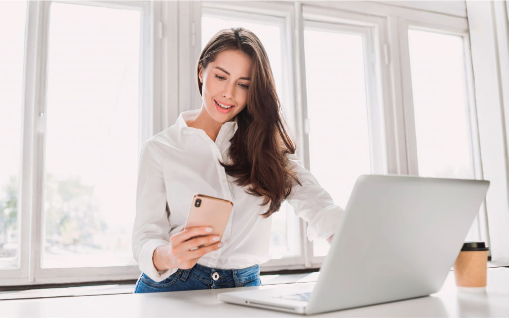 mulher branca segurando celular na mão direita sentada diante de computador e pesquisando a alíquota do imposto de renda sobre bônus