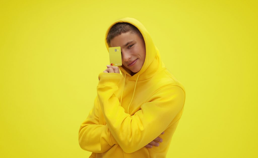 Foto de um garoto com edredon amarelo segurando um cartão também amarelo da conta C6 Yellow num fundo amarelo.