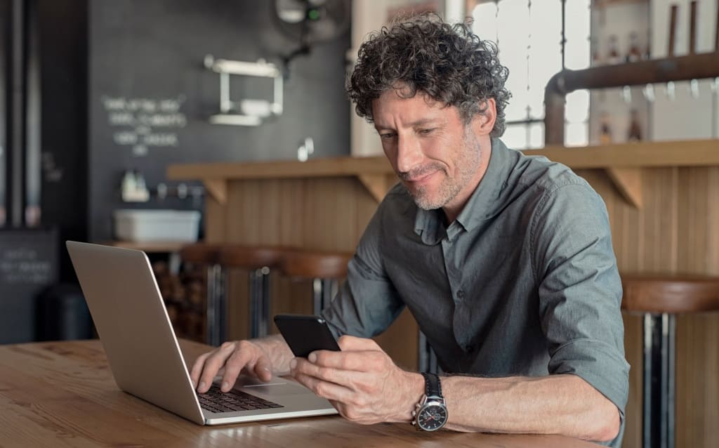 homem grisalho com roupa social sorrindo sentado diante de computador segurando celular nas mãos e pesquisando o que é o preço justo de uma ação