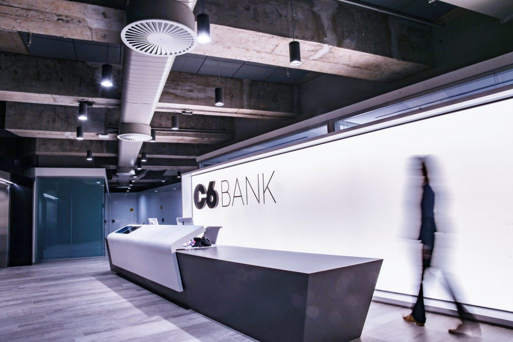 Foto da recepção do 7º andar do prédio do C6 Bank, onde se pode ver a bancada cinza da recepção, a frente de um painel branco com o  logo do C6 Bank ao fundo. Na imagem, também é possível ver uma pessoa em movimento.