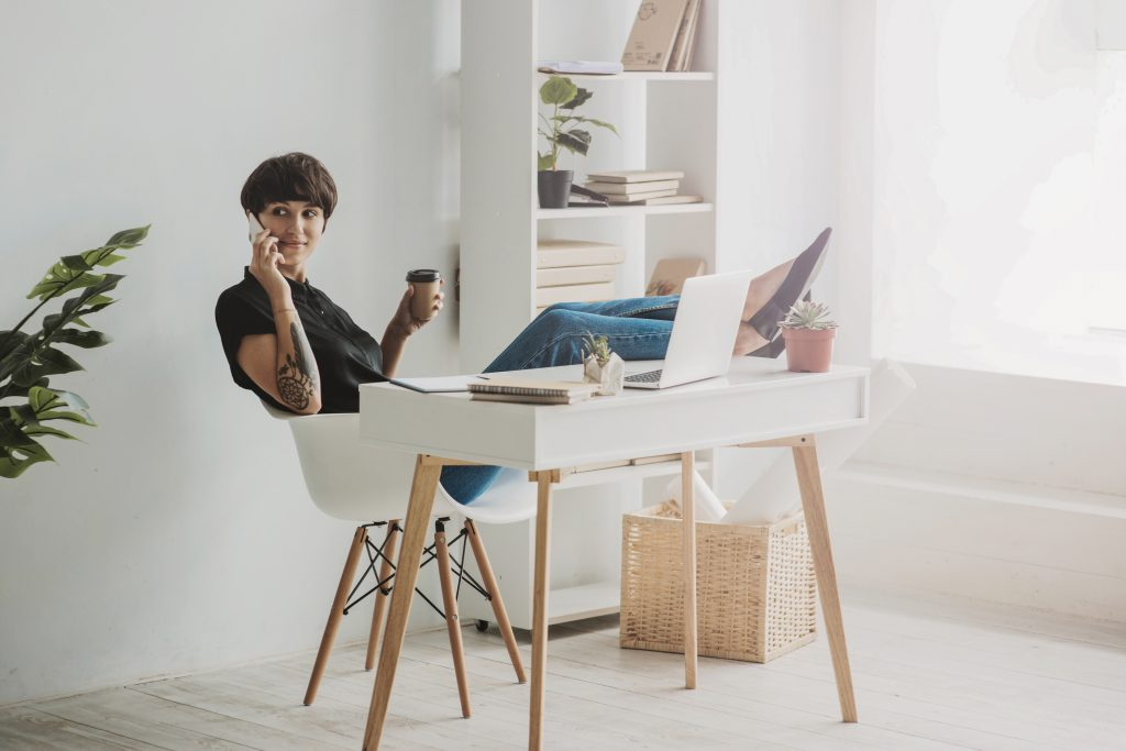 Foto de uma jovem branca, morena, com o cabelo curto no estilo channel. Ela está sentada numa cadeira com os dois pés apoiados em cima de uma mesa a sua frente, segurando um celular na orelha com a mão direita e um café com a mão esquerda. Na cena ainda vemos um notebook e alguns papéis em cima da mesa e uma estante ao fundo.
