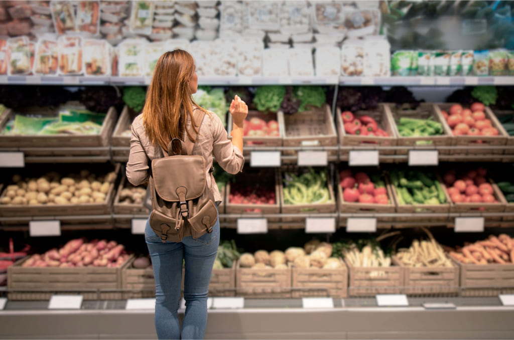 mulher branca parada diante de várias frutas e vegetais em um mercado, produtos que estão entre as principais commodities do brasil