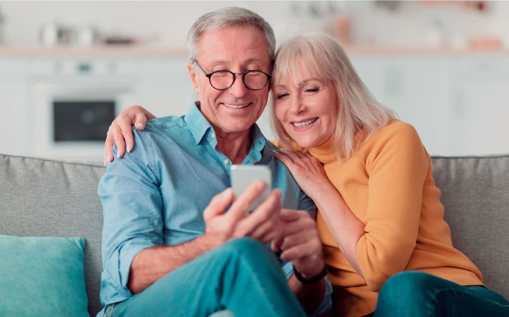 homem e mulher brancos e idosos sorriem sentados em sofá enquanto olham para celular após aprenderem o que é progressiva compensável