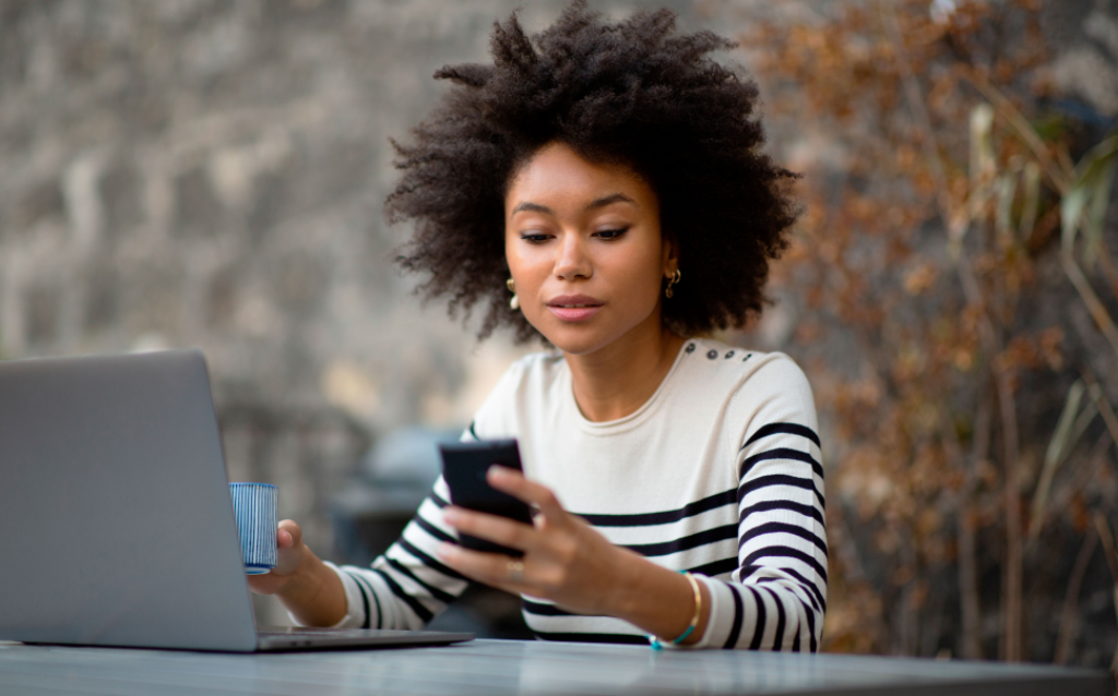Mulher negra com blusa listrada, segurando o celular em frente ao computador pesquisando a diferença entre crv e crlv
