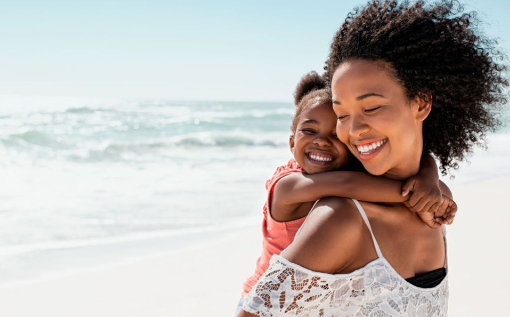 mãe negra de cabelos cacheados carregando filha nas costas à beira do mar durante o ano sabático