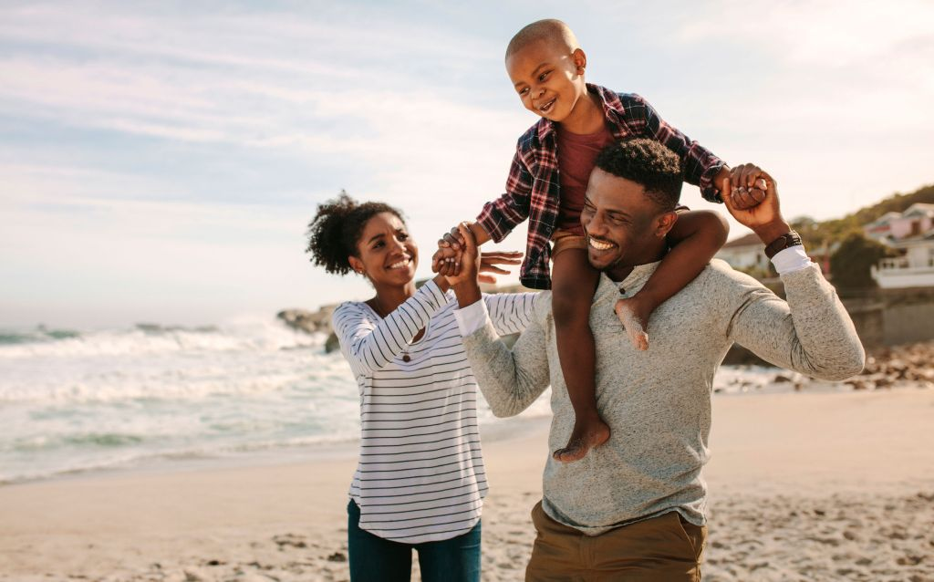 a imagem apresenta três pessoas na praia que parecem formar uma família, um casal com seu filho onde a mãe é uma mulher negra e está sorrindo enquanto observa o filho pequeno, que está sentado nos ombros do pai, que sorri enquanto caminha e segura a criança