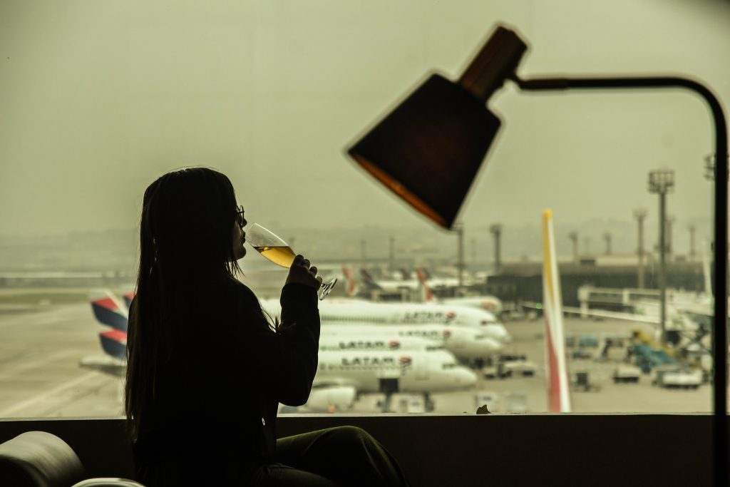 Uma pessoa sentada em um lounge de aeroporto, segurando uma taça de espumante enquanto observa a pista. Ao fundo, aviões da LATAM estacionados e estruturas de iluminação do aeroporto, com um ambiente de iluminação suave e uma luminária visível no canto superior direito.