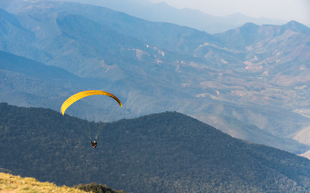 Praticante de parapente voando sobre montanhas verdejantes, em uma paisagem deslumbrante e cheia de natureza. O céu azul complementa a cena.
