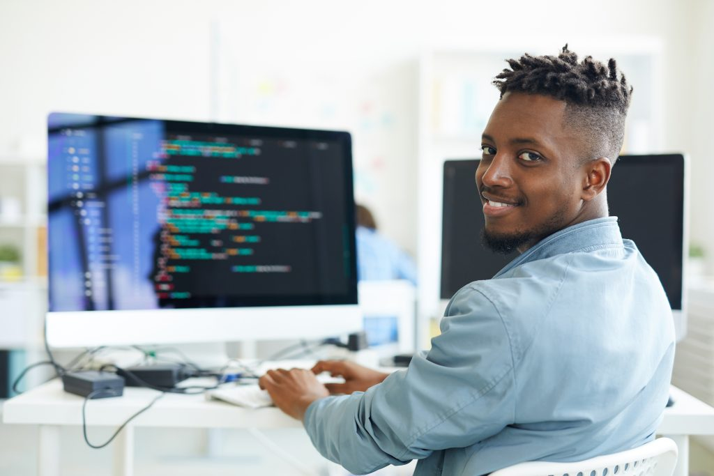 Homem negro sentando em frente ao computador olhando para trás e sorrindo, pensando em seu perfil de investidor