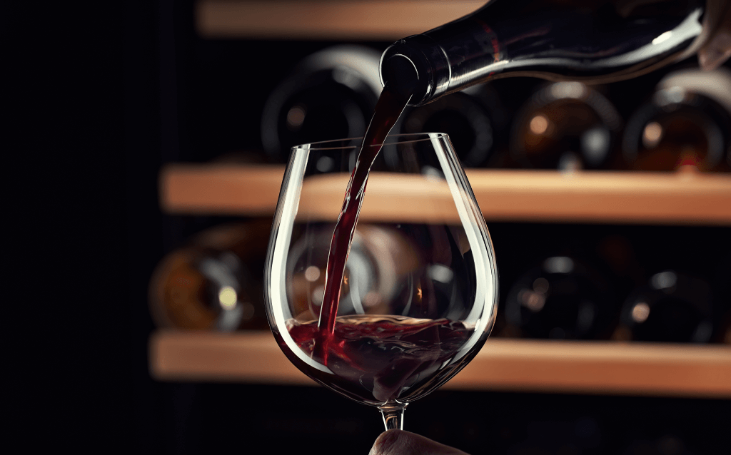 Imagem de um vinho tinto sendo derramado em uma taça, destacando sua cor vibrante. A cena é iluminada com um fundo de adega.