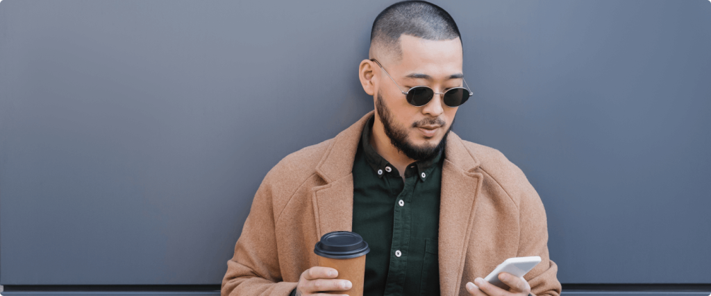 Imagem mostra homem de cabelo curto, barba e óculos escuros encostado em uma parede. Com uma das mãos ele segura um copo de café e, com a outra, segura um celular. Ele veste camisa verde com um smoking marrom.
