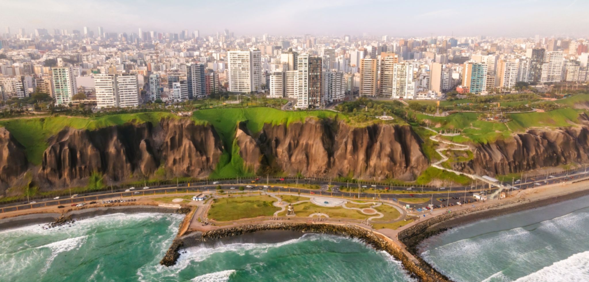 Mostra a cidade de Lima vista do alto, com falésias extensas cobertas por vegetação verde na parte central. Abaixo, há uma avenida costeira e o mar com ondas próximas à costa. Ao fundo, aparecem diversos prédios residenciais e comerciais compondo o skyline urbano.