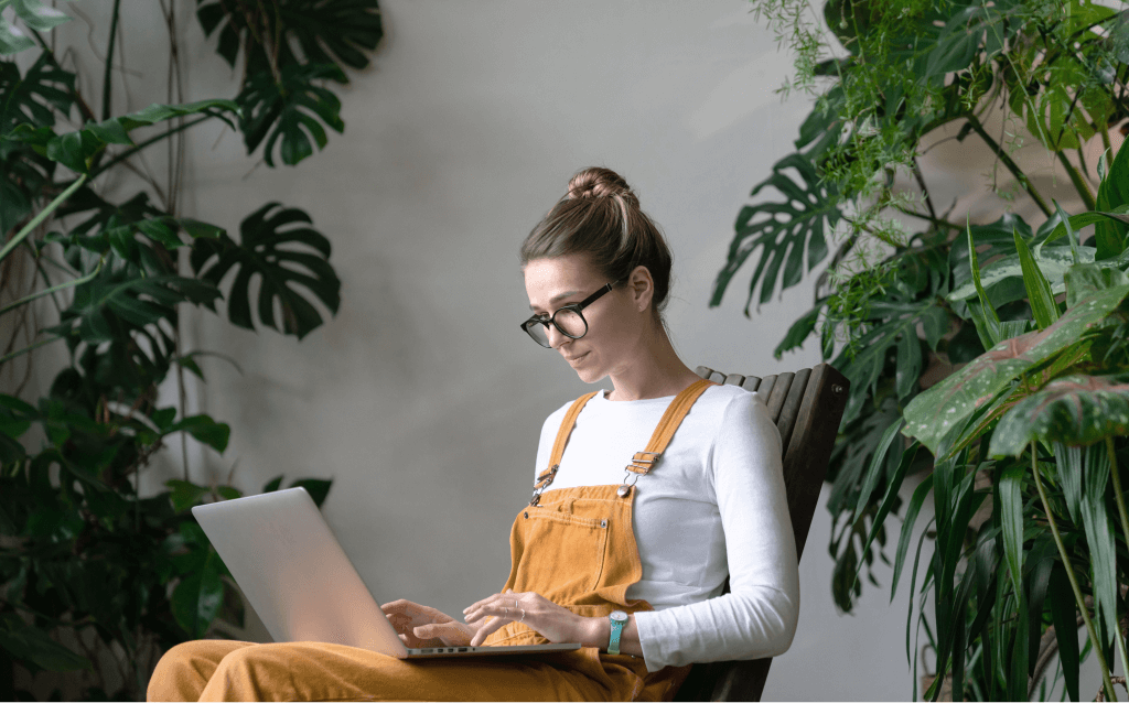 Mulher jovem usando óculos e macacão mostarda, sentada em uma cadeira de madeira, trabalhando em um notebook cercada por plantas tropicais.