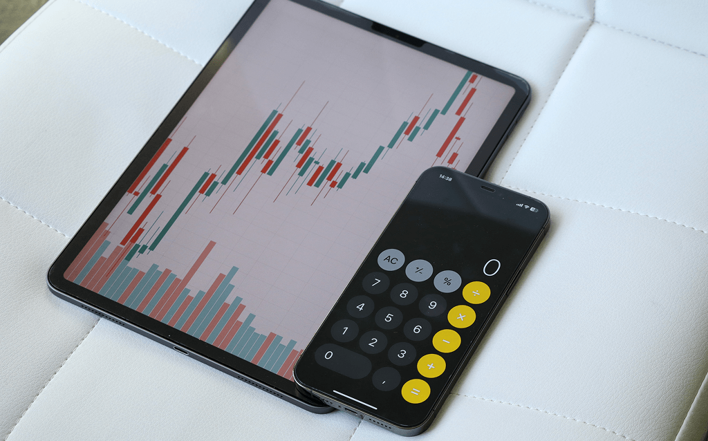 Tablet com gráfico de velas de ações e barras verticais ao lado de um smartphone exibindo uma calculadora digital com o número zero na tela, ambos apoiados sobre um assento branco.
