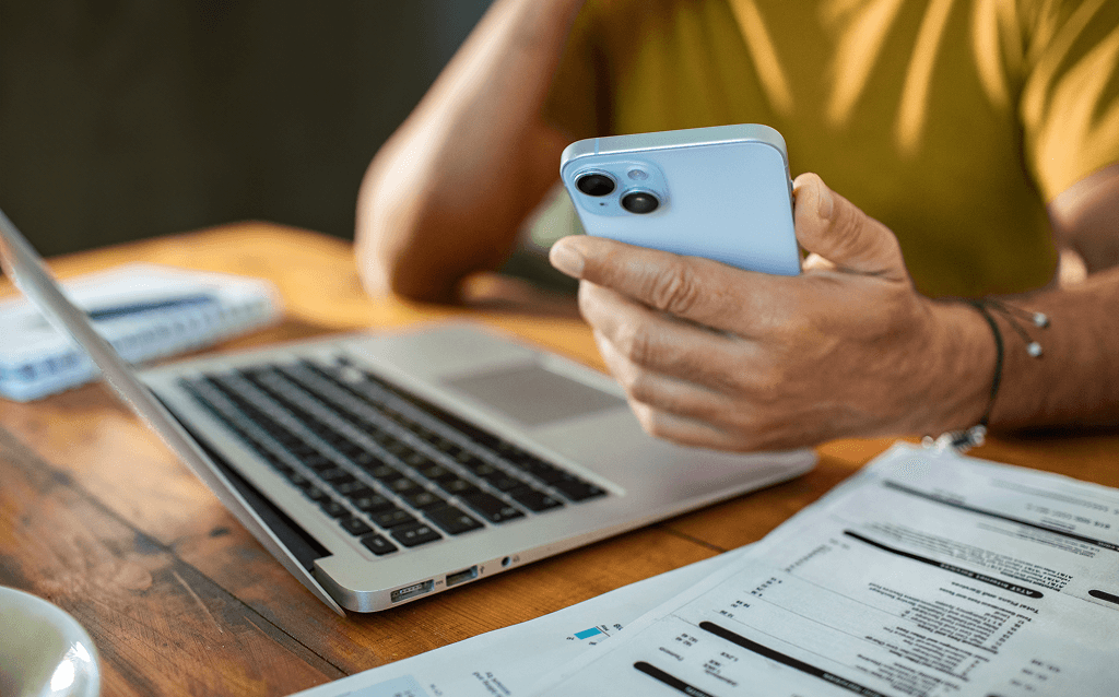 Pessoa usando um smartphone enquanto trabalha em um notebook, com papéis de contas e faturas sobre a mesa. A cena sugere uma atividade de organização financeira ou pagamento de contas.