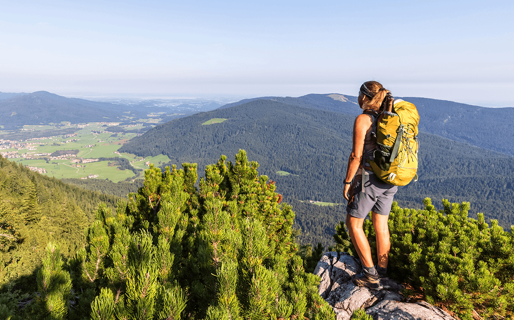 Pessoa com mochila amarela faz trilha em região montanhosa, parada sobre uma pedra, observando uma vista ampla de vales verdes, florestas e vilarejos ao fundo. O dia está claro e ensolarado.