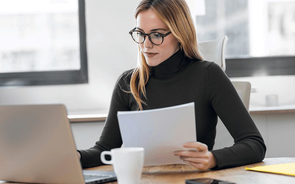 Mulher com óculos e blusa de gola alta preta sentada em frente a um notebook, segurando folhas de papel e olhando atentamente para a tela. Ela está em um ambiente de escritório, com uma xícara branca sobre a mesa.