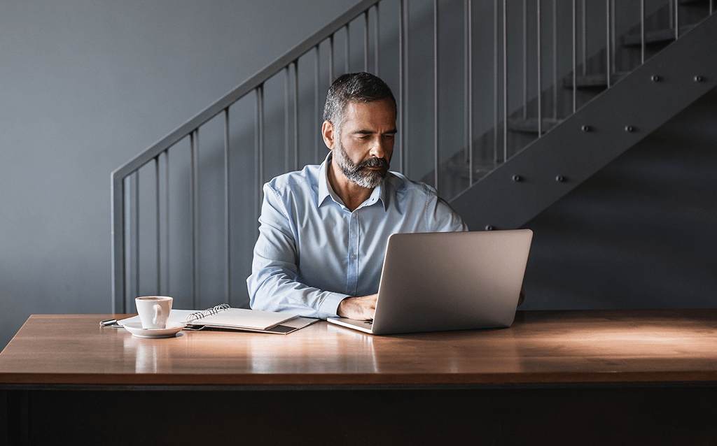 Homem de meia-idade com barba grisalha usando camisa azul clara, sentado à mesa em frente a um notebook, com um caderno e uma xícara de café ao lado, em ambiente moderno com escada metálica ao fundo.